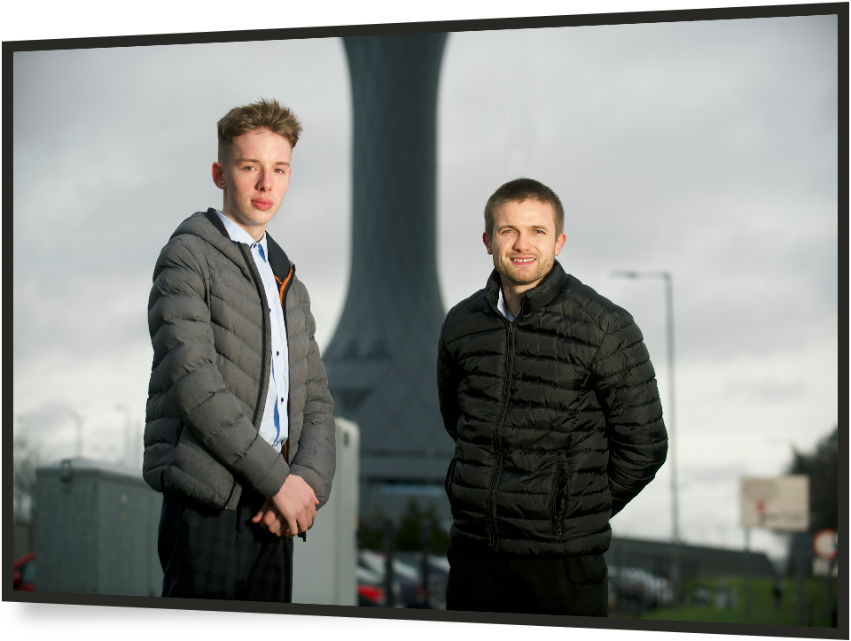 a young male and older male looking at the camera in front of a power station