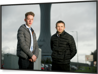 a young male and older male looking at the camera in front of a power station